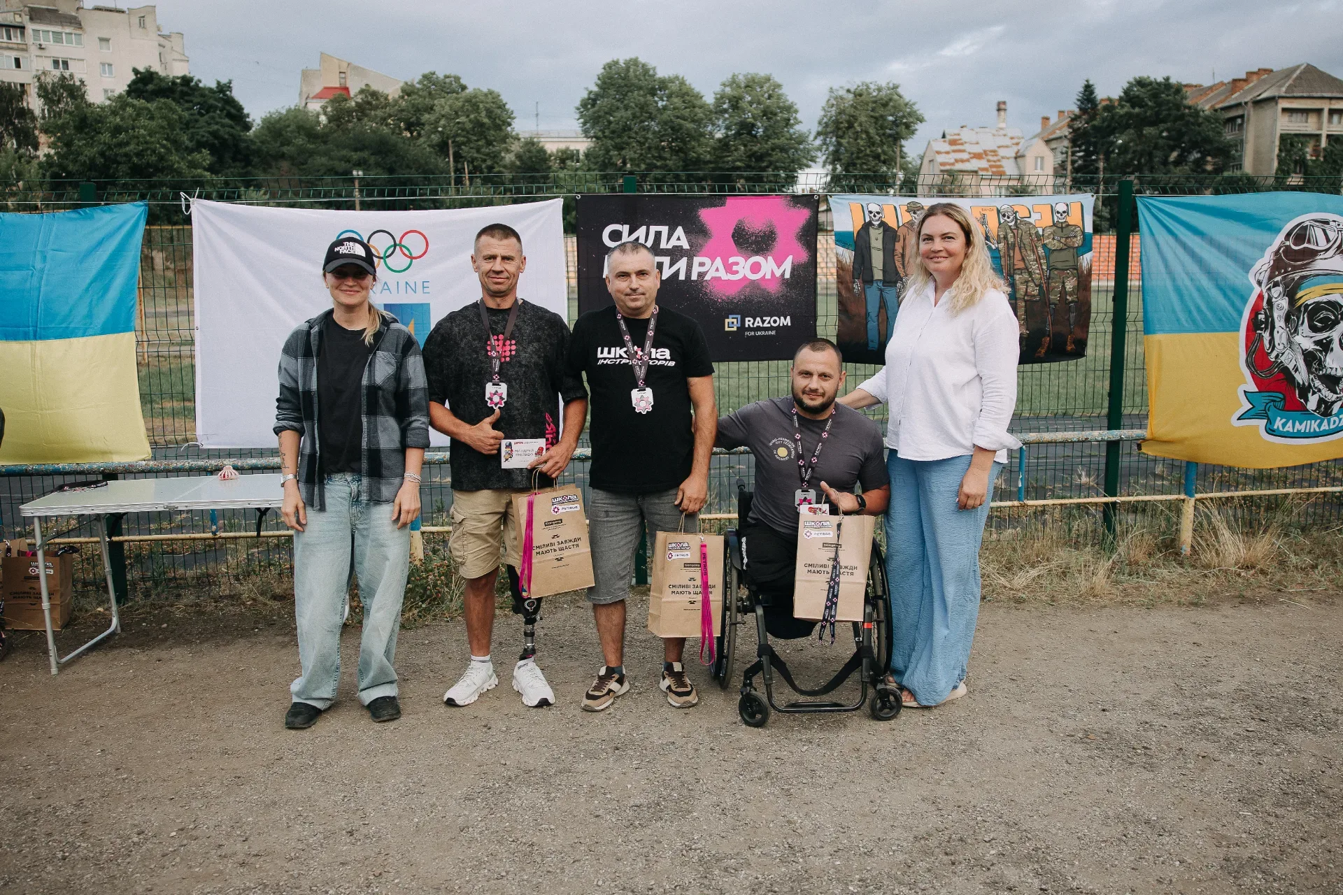 Кубок Петроса 2026 На фото п’ятеро людей позують після заходу. Троє чоловіків у центрі тримають подарункові пакети та нагороди, один із них має протез ноги, інший сидить у кріслі колісному. По боках стоять дві жінки. Позаду розміщені українські прапори, банери з олімпійською символікою та написом «Сила в єдності разом». Зйомка відбувається просто неба на спортивному майданчику.